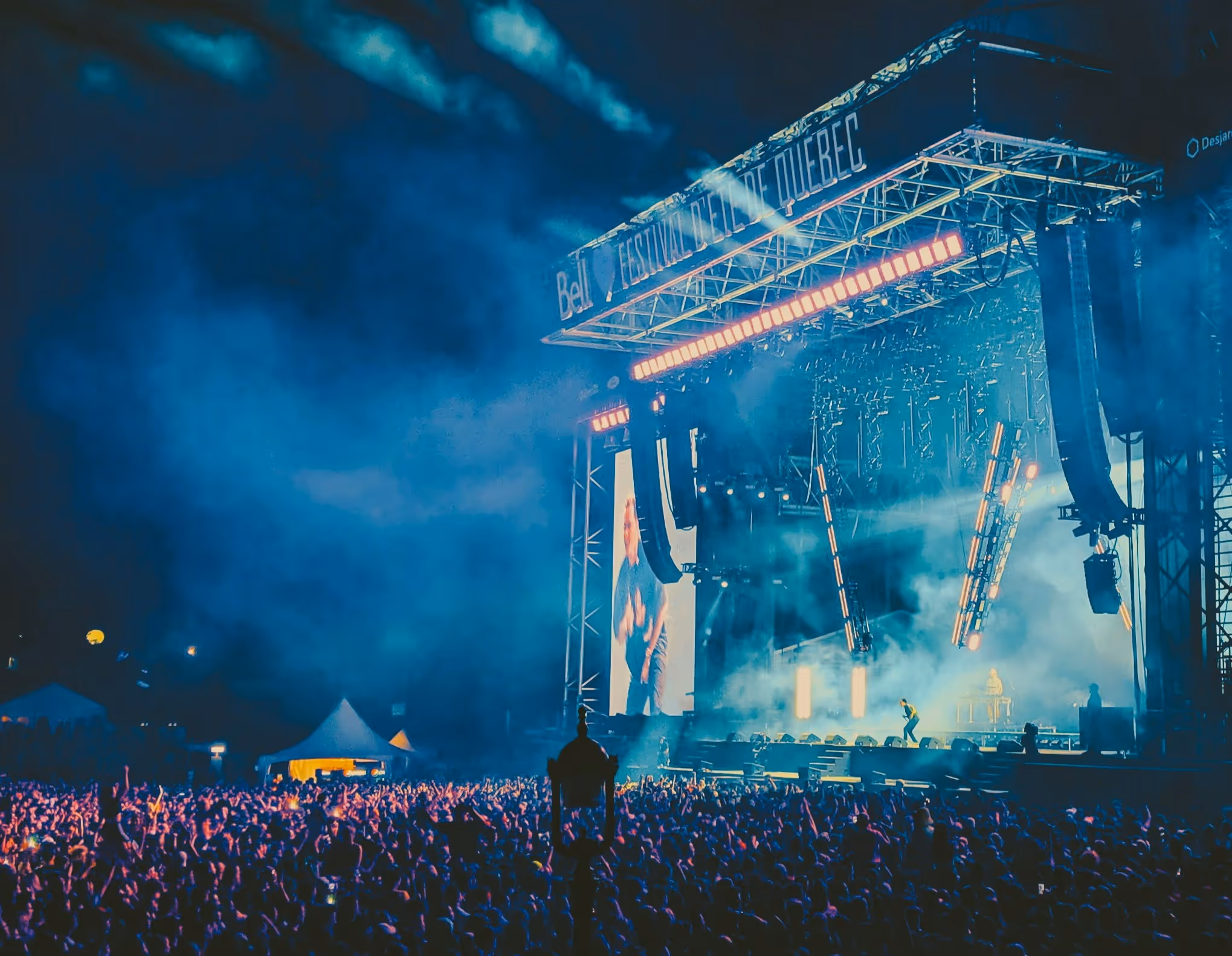 Large crowd enjoying a nighttime outdoor concert at Festival d'été de Québec, with bright stage lights, smoke effects, and dynamic visuals on big screens.